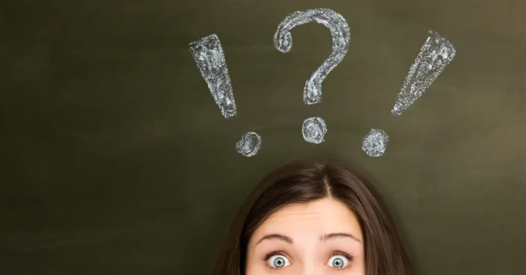 Rosto de uma jovem com olhos arregalados, expressando surpresa ou dúvida, com pontos de exclamação e interrogação desenhados acima da cabeça em um quadro-negro.