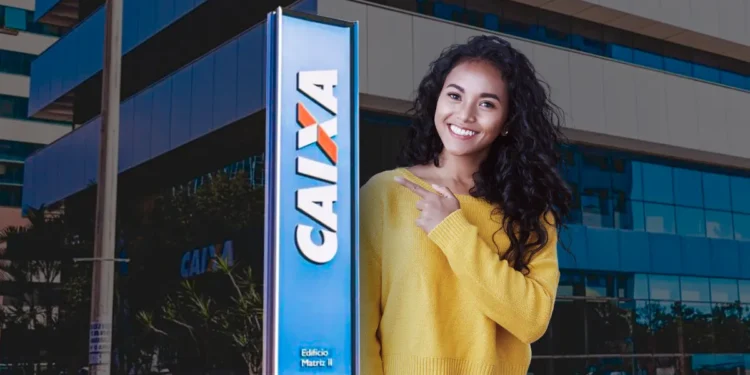 Jovem mulher sorridente com blusa amarela apontando para placa azul da Caixa Econômica em frente a prédio corporativo.