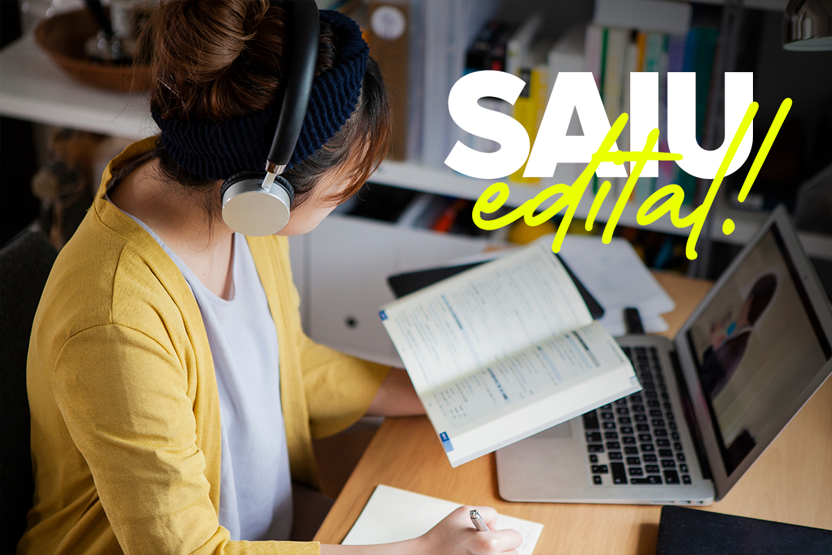 Mulher usando fones de ouvido estudando com livro aberto e anotando em caderno ao lado de laptop.