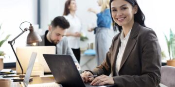 Mulher sorrindo enquanto trabalha em um notebook em um escritório moderno, com colegas ao fundo, representando as futuras profissões.