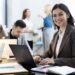 Mulher sorrindo enquanto trabalha em um notebook em um escritório moderno, com colegas ao fundo, representando as futuras profissões.