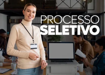 Mulher sorridente com crachá em ambiente de escritório durante processo seletivo com laptop e pessoas ao fundo.