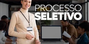 Mulher sorridente com crachá em ambiente de escritório durante processo seletivo com laptop e pessoas ao fundo.