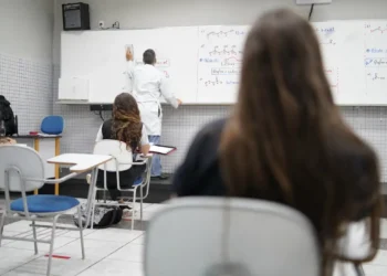 Sala de aula com alunos sentados em carteiras assistindo professor escrevendo fórmulas químicas no quadro branco.
