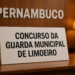 Placa com os dizeres 'Concurso da Guarda Municipal de Limoeiro' sobre uma mesa de madeira.