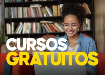 Jovem sorridente usando notebook em sofá com estante de livros ao fundo.