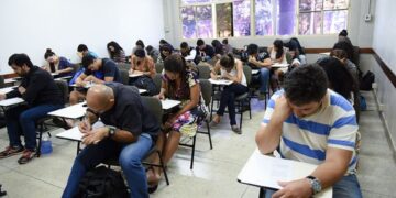 Grupo de pessoas sentadas em carteiras individuais escrevendo durante aplicação de prova.