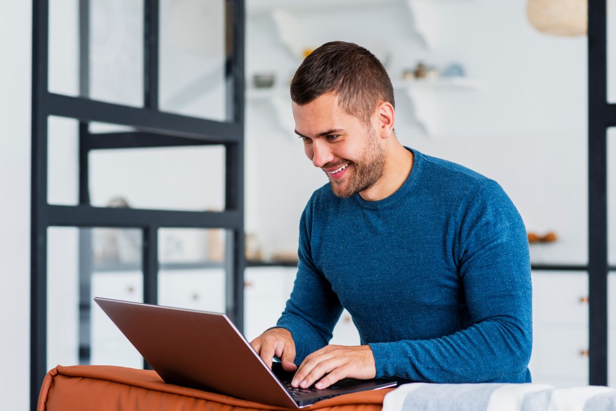 Inscrição concurso PM AL Homem sorridente usando notebook em sala iluminada com decoração contemporânea.