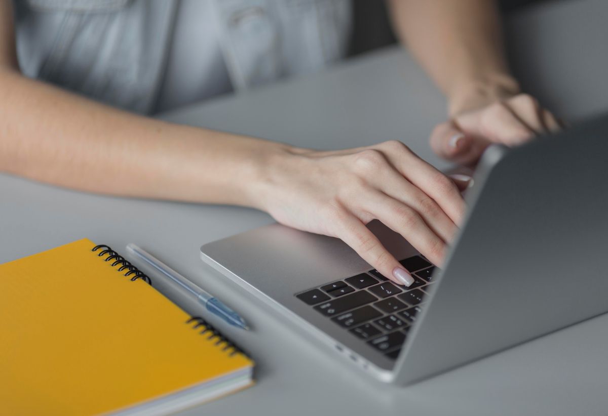 Inscrição em concurso 2026. Mãos femininas digitando em notebook com caderno amarelo e caneta ao lado.