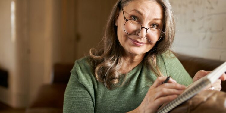 Mulher de óculos anotando em caderno enquanto sorri sentada em sofá.