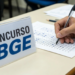Candidato preenche cartão-resposta com caneta ao lado de placa do concurso IBGE