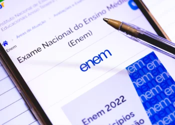 Close-up de um smartphone exibindo a página oficial do Enem no portal do Inep, com uma caneta esferográfica sobre a tela e folhas de caderno ao fundo.