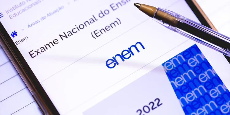 Close-up de um smartphone exibindo a página oficial do Enem no portal do Inep, com uma caneta esferográfica sobre a tela e folhas de caderno ao fundo.