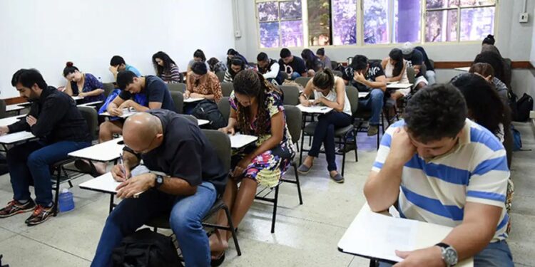 aplicação prova encceja sala de aula quatro provas redação Sala de aula cheia de jovens e adultos sentados em carteiras, concentrados resolvendo a prova impressa do Encceja, com um aplicador visível ao fundo.