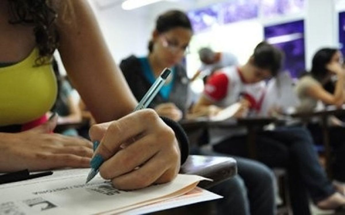 O que muda no Enem 2026 para o estudante com o novo papel do exame Estudante escrevendo em folha de prova em sala de aula com outros candidatos ao fundo — o que muda no Enem 2026 para o estudante