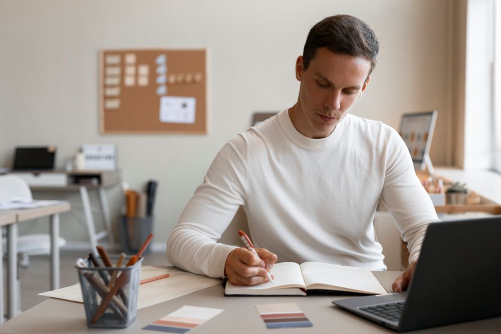 estudante organizando cronograma estudos enem Close-up de um homem em home office escrevendo em um caderno aberto, com um laptop ligado e paletas de cores sobre a mesa, concentrado no planejamento.