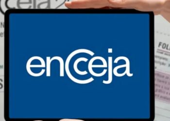 Pessoa segurando um tablet que mostra o logotipo do Encceja em branco sobre fundo azul, com folhas impressas da prova de redação desfocadas ao fundo.