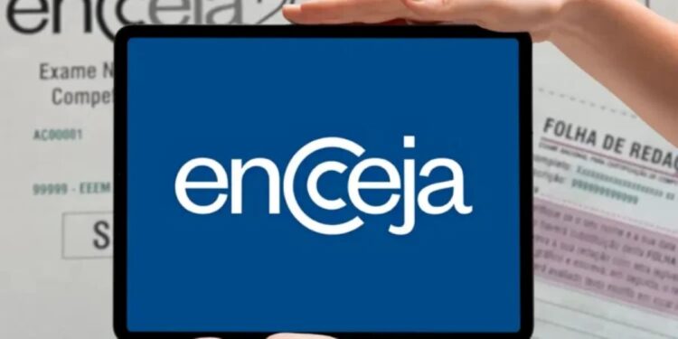 Pessoa segurando um tablet que mostra o logotipo do Encceja em branco sobre fundo azul, com folhas impressas da prova de redação desfocadas ao fundo.
