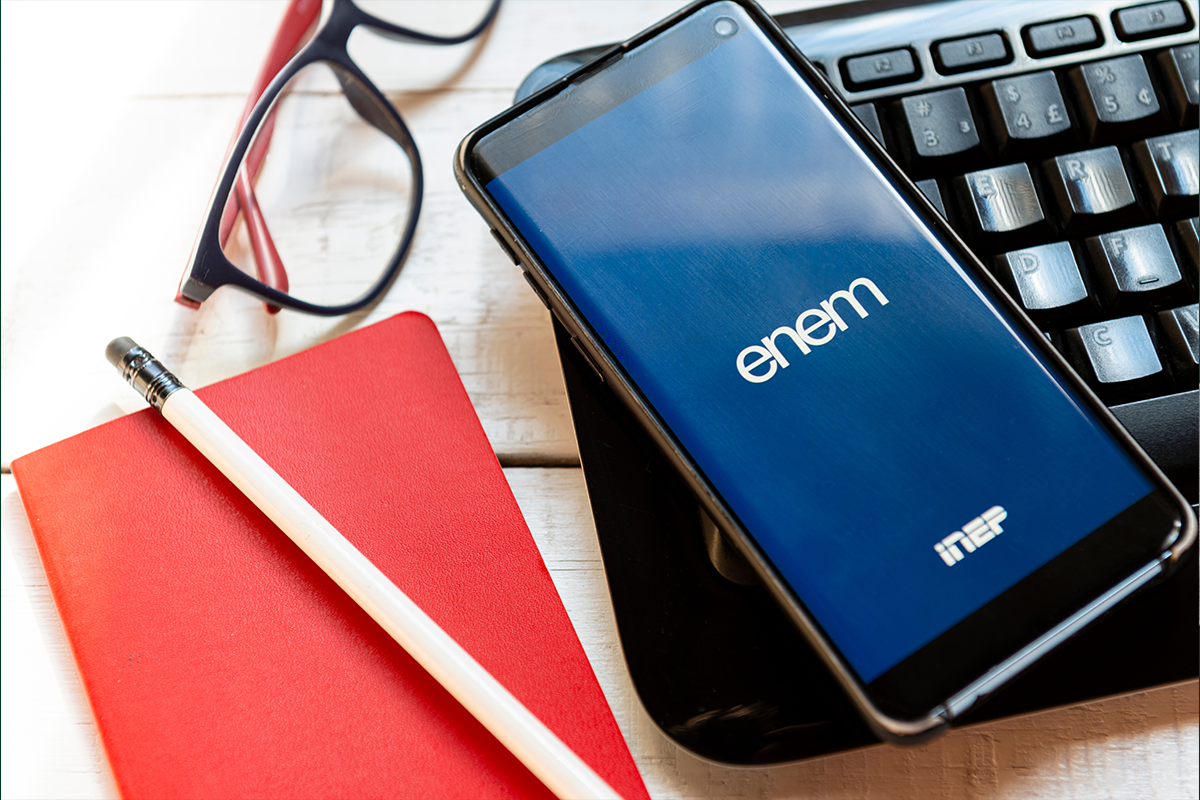 o que esperar redação enem 2026 estrutura Uma mesa branca de estudo vista de cima com um smartphone exibindo o logotipo 'enem' e 'INEP' sobre um notebook, ao lado de um caderno vermelho, lápis e óculos.