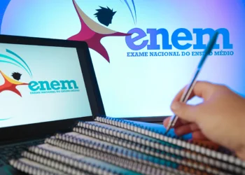 Close de uma mão segurando uma caneta sobre cadernos de espiral, com um notebook e um monitor ao fundo exibindo o logotipo oficial do Enem.
