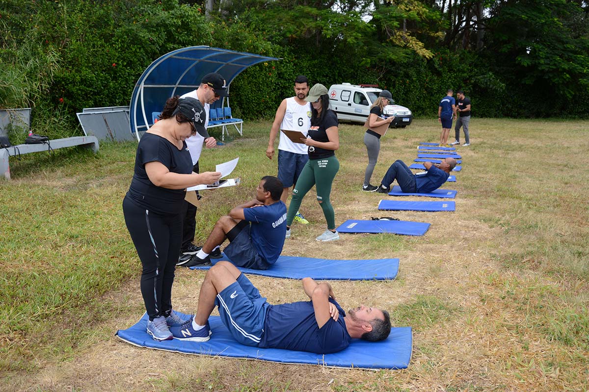 TAF Guarda Municipal Candidatos realizando abdominais em colchonetes, avaliadores conferem pranchas e preenchem formulários