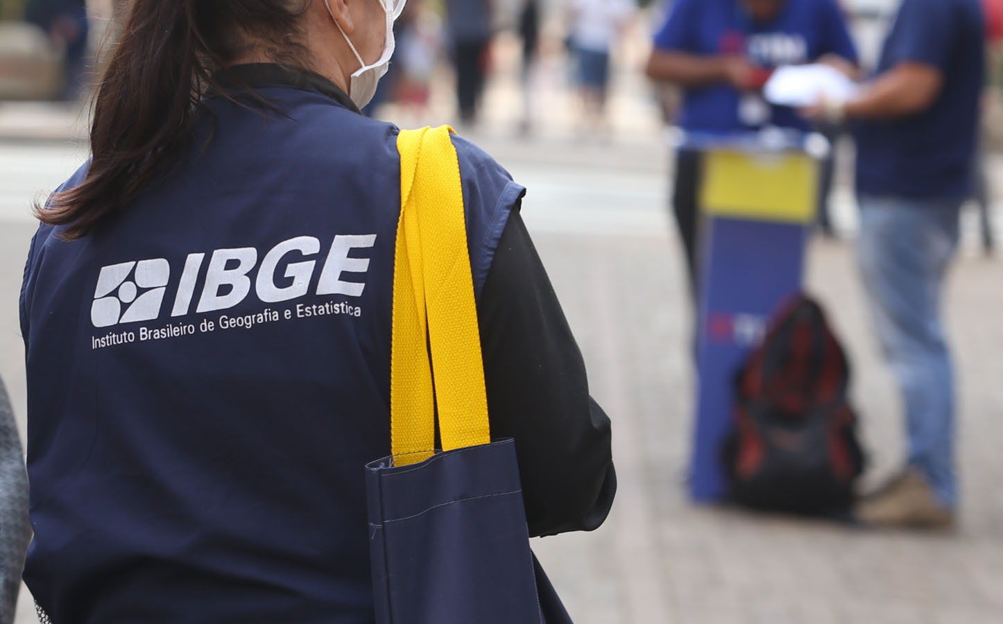Vagas concurso IBGE 2026 Agente do IBGE de colete azul e máscara segura bolsa amarela em rua movimentada