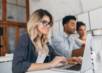 Jovem loira de óculos, vestindo camisa azul sobre blusa branca, sorrindo levemente enquanto digita em um notebook em uma mesa branca. Ao fundo, borrados, dois colegas (um homem negro de camisa xadrez e uma mulher negra) também trabalham em notebooks, ilustrando um ambiente de trabalho jovem e colaborativo.