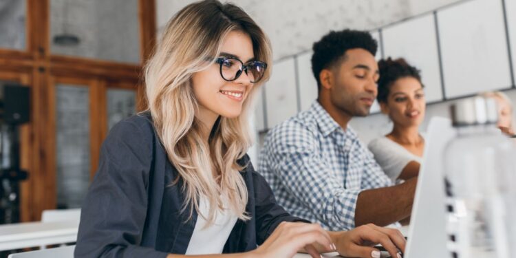 Jovem loira de óculos, vestindo camisa azul sobre blusa branca, sorrindo levemente enquanto digita em um notebook em uma mesa branca. Ao fundo, borrados, dois colegas (um homem negro de camisa xadrez e uma mulher negra) também trabalham em notebooks, ilustrando um ambiente de trabalho jovem e colaborativo.