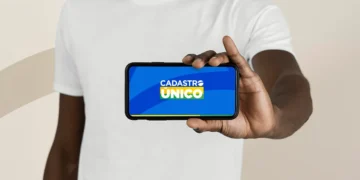 Homem negro de camiseta branca segura smartphone horizontal com tela do aplicativo Cadastro Único para Programas Sociais do Governo Federal