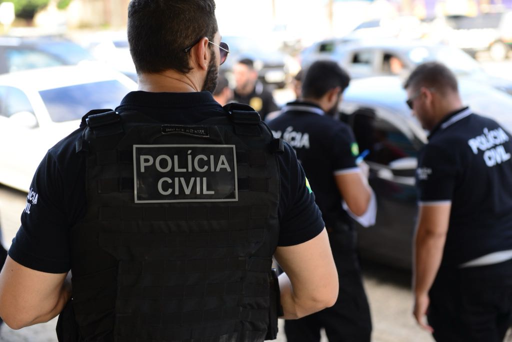 Concurso PC TO Três agentes da Polícia Civil trabalham na rua ao lado de veículos, um de colete na frente