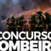 Bombeiros com equipamentos de proteção e cilindros de oxigênio direcionam mangueiras contra chamas em cenário de incêndio