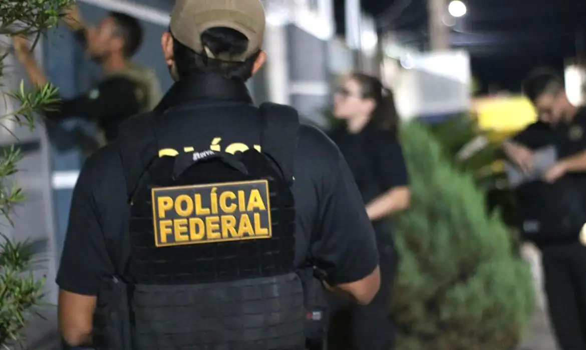 Concurso PF Agente da Polícia Federal de colete preto e boné bege observa ação policial ao ar livre à noite