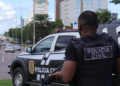 Policial Civil de colete tático segura arma próximo a viatura em avenida urbana movimentada