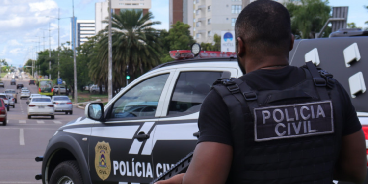 Policial Civil de colete tático segura arma próximo a viatura em avenida urbana movimentada