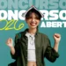 Jovem sorridente com livro aberto na cabeça, jaqueta verde e óculos pendurados, celebrando concursos 2026