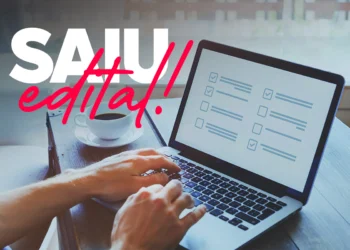 Mãos digitando em notebook prateado exibindo lista de tarefas com checkboxes, xícara de café ao lado, texto "SAIU edital!" sobreposto