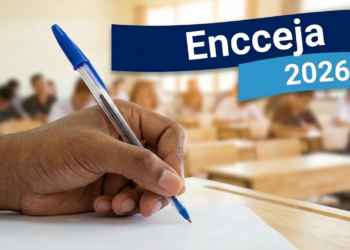 Mão segurando caneta azul escreve em papel com textos "Encceja" e "2026" em destaque, sala de aula ao fundo