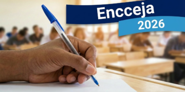 Mão segurando caneta azul escreve em papel com textos "Encceja" e "2026" em destaque, sala de aula ao fundo