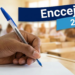 Mão segurando caneta azul escreve em papel com textos "Encceja" e "2026" em destaque, sala de aula ao fundo