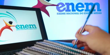 Mão segurando caneta sobre cadernos empilhados diante de telas com logo do Enem em fundo azul