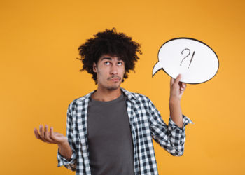 Homem jovem com cabelo afro segura balão de fala branco com símbolos ?! enquanto gesticula dúvida em fundo amarelo
