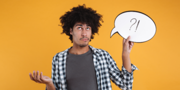 Homem jovem com cabelo afro segura balão de fala branco com símbolos ?! enquanto gesticula dúvida em fundo amarelo