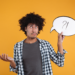 Homem jovem com cabelo afro segura balão de fala branco com símbolos ?! enquanto gesticula dúvida em fundo amarelo