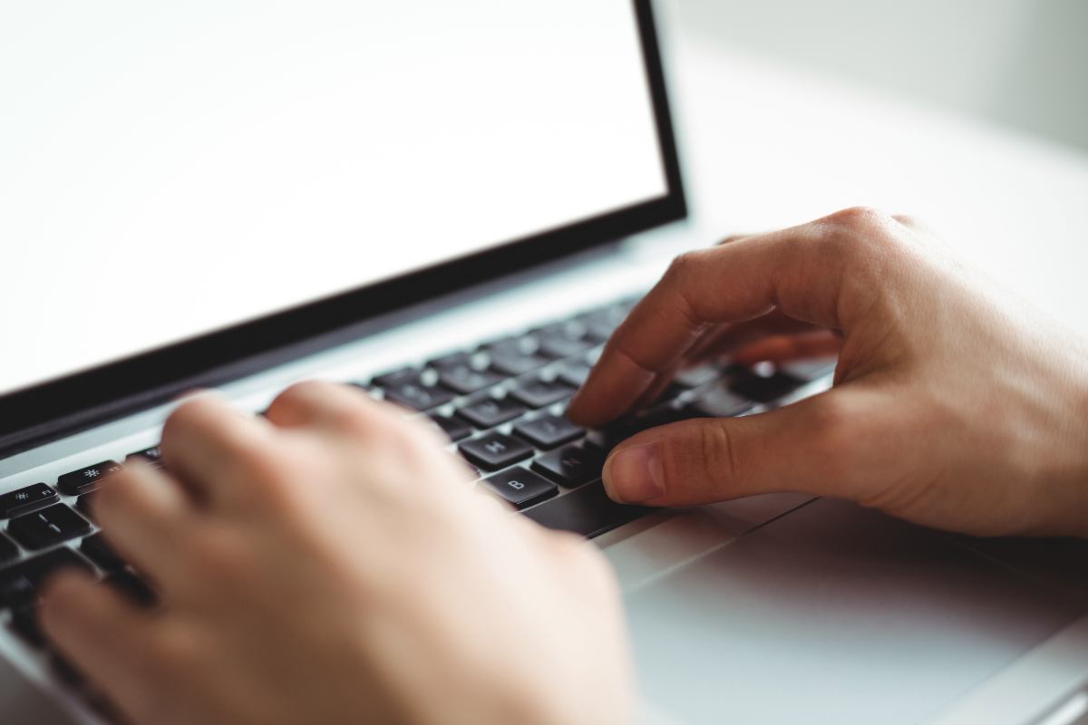 Inscrições Concurso Duas mãos digitando em teclado preto de notebook prateado com tela iluminada em branco ao fundo