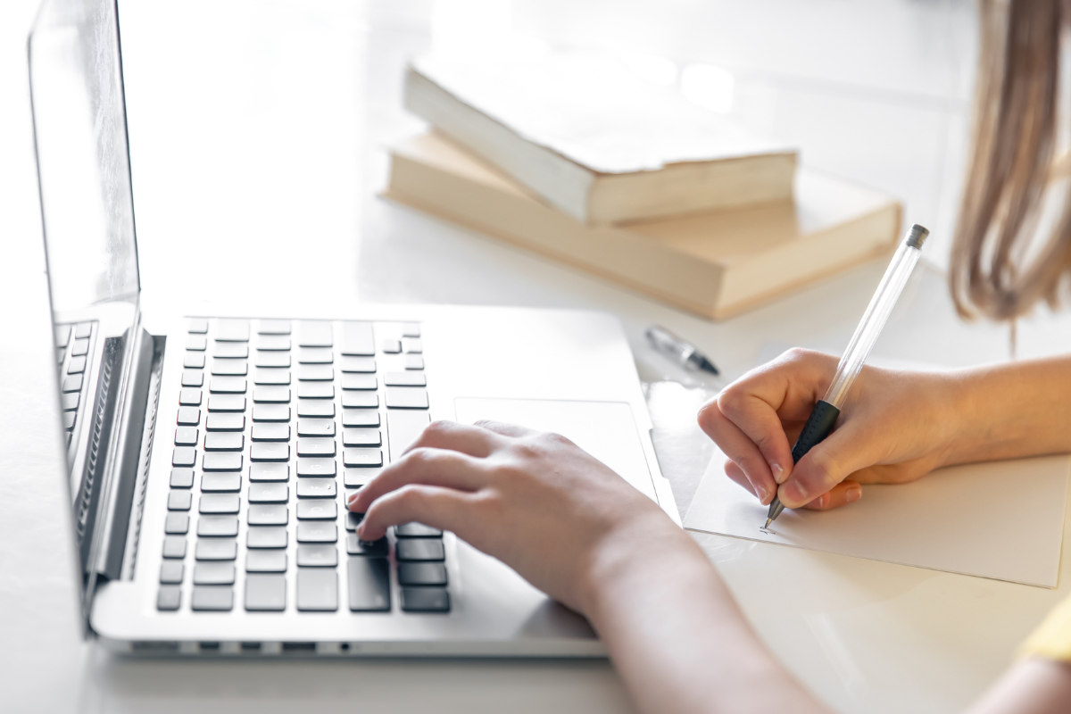 Inscrição Concurso Mãos femininas digitando em laptop prateado enquanto escrevem em papel com caneta branca, livros ao fundo