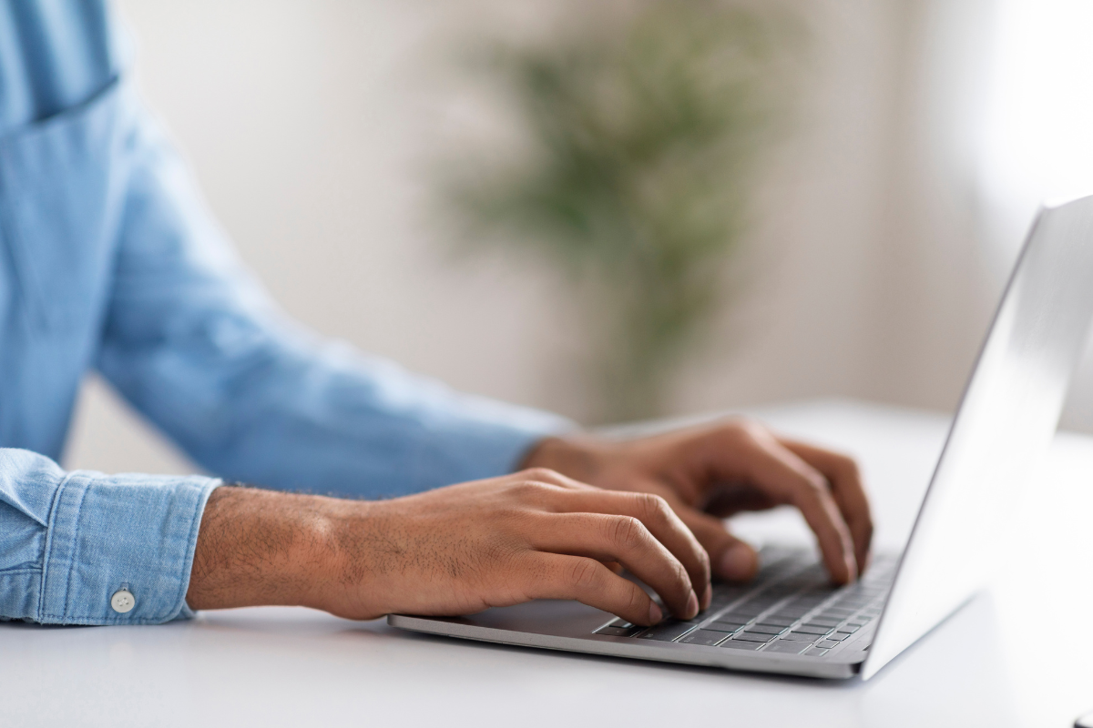 Inscrição Prefeitura Mãos de homem com camisa jeans azul digitando em notebook cinza prata sobre mesa branca com planta ao fundo