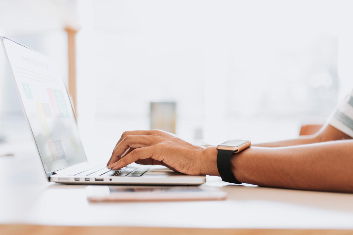 Inscrição Prefeitura Mãos digitando em laptop prateado com smartwatch preto, smartphone rosé ao lado sobre mesa de madeira clara