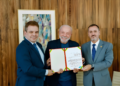 Presidente Lula entre dois homens de terno segurando documento com fitas verde-amarela e vermelha no Planalto