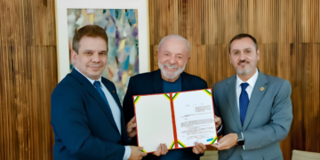 Presidente Lula entre dois homens de terno segurando documento com fitas verde-amarela e vermelha no Planalto