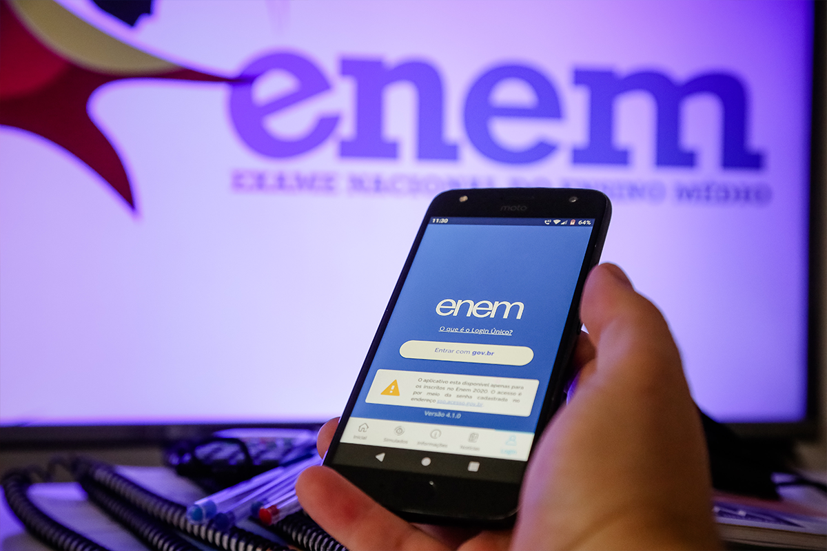 Prazo justificativa Enem Mão segurando smartphone Motorola exibindo tela de login do app ENEM com opção "Entrar com gov.br" e monitor ao fundo